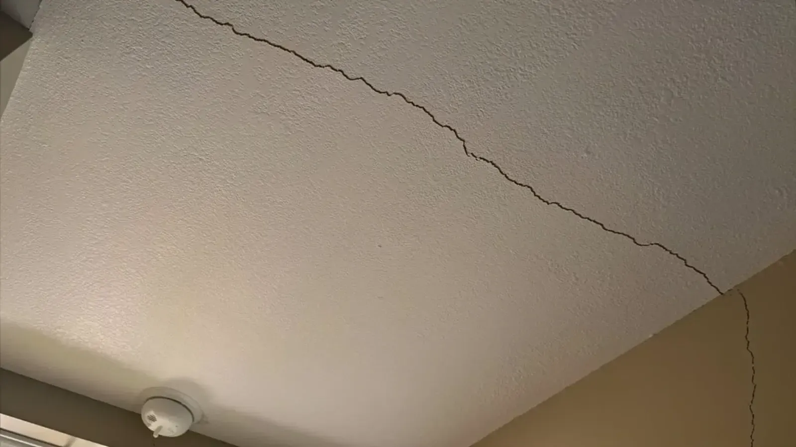 A long crack running diagonally across a lightly textured painted ceiling toward the corner where the ceiling meets the wall, with a round white smoke detector mounted nearby