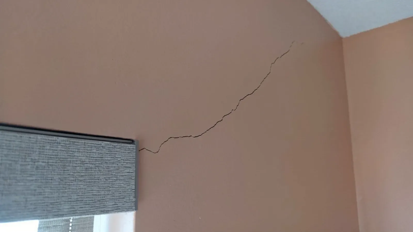 A hairline stress crack running diagonally across a painted interior wall from the head of a window casing up toward the wall-and-ceiling corner