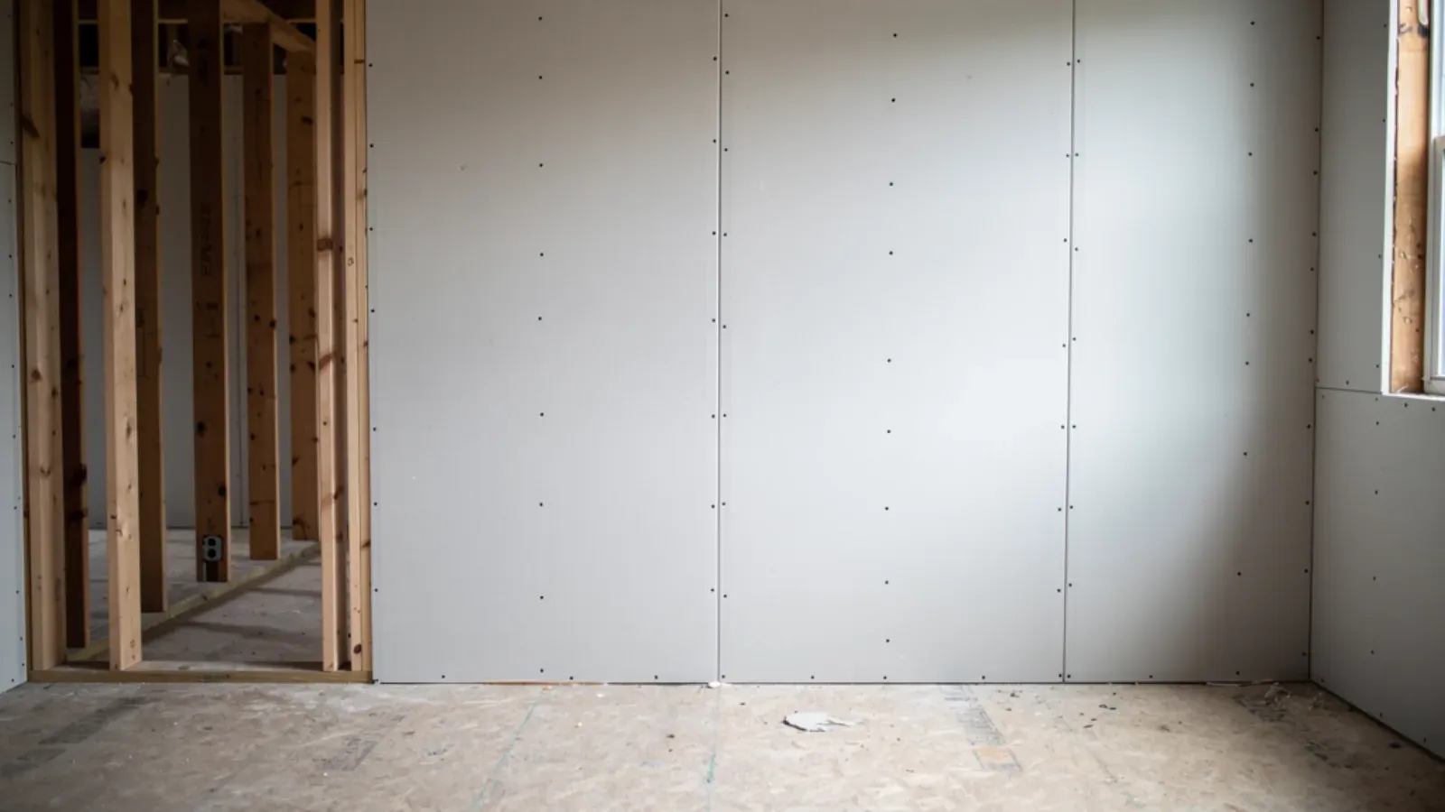 Bare gypsum drywall panels screwed onto wood wall studs in a room under construction, one stud bay still open