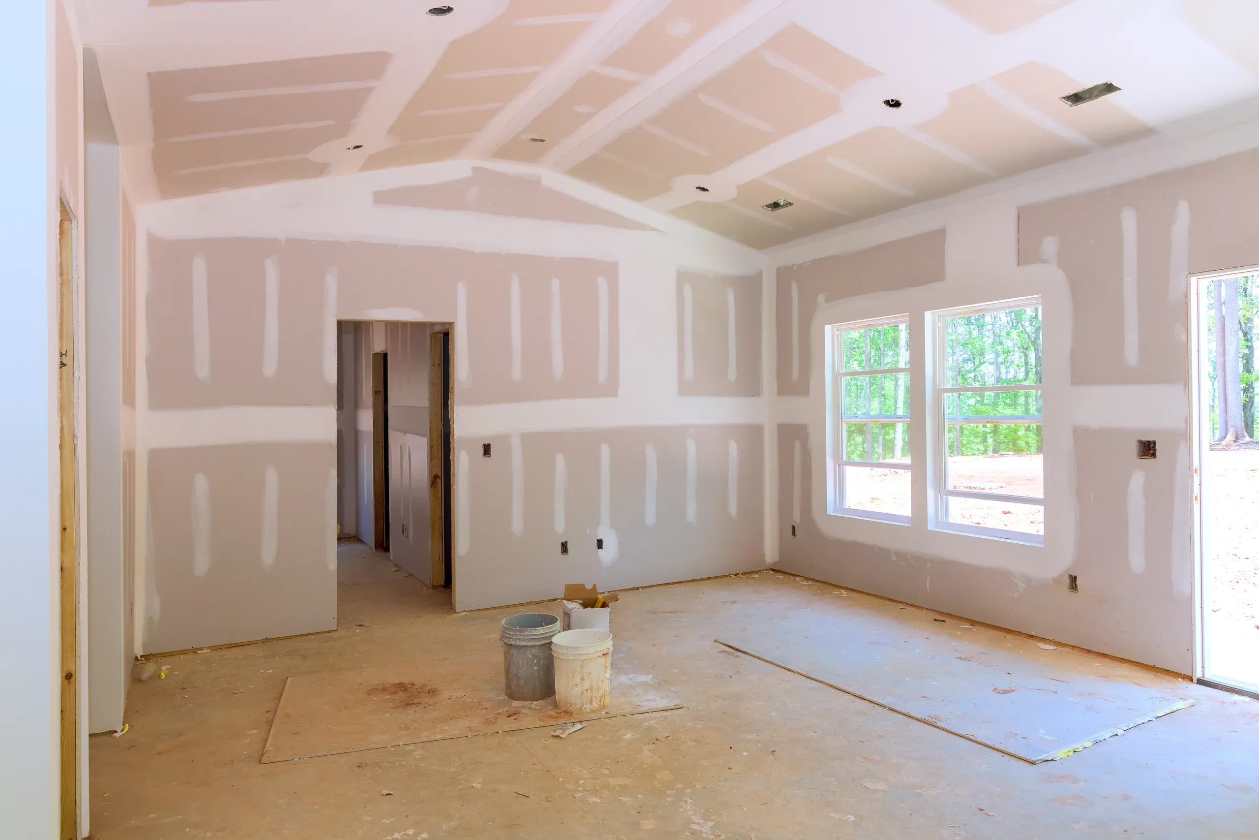 Newly hung drywall room with every seam taped and coated in white joint compound and each fastener spotted, ceiling and walls unsanded, two compound buckets on the bare subfloor