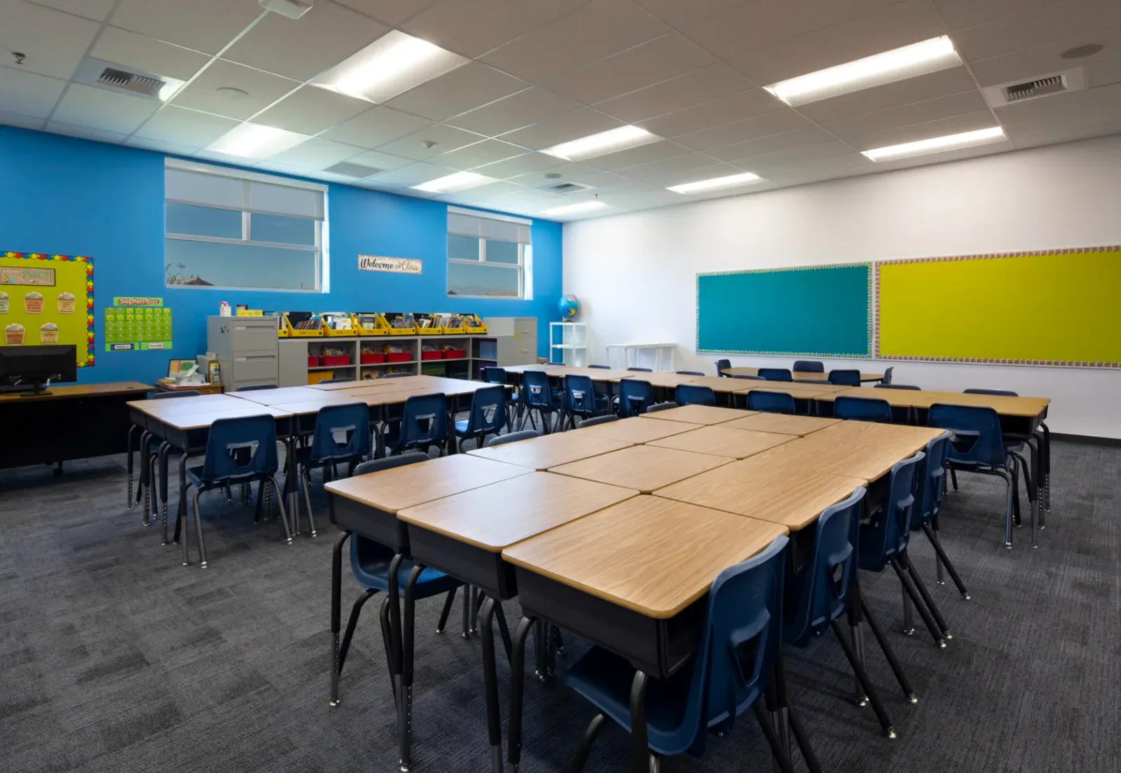 Classroom with finished drywall walls in blue and white, a suspended acoustic-tile ceiling, tackboards, and clustered student desks with chairs on carpet tile