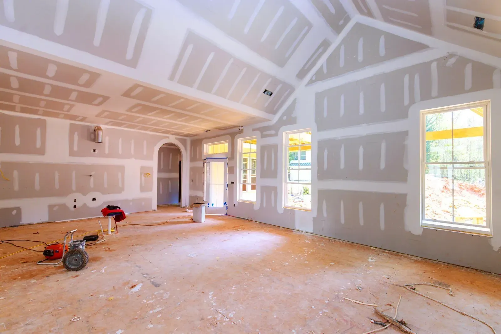 Bright vaulted great room under construction with freshly hung and taped drywall on the walls and sloped ceiling, three tall windows, and tools on the plywood subfloor
