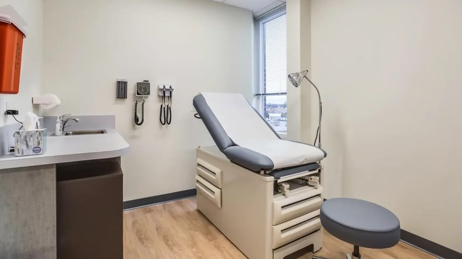 A medical exam room with an exam table against a broad plain wall, a sink counter and wall-mounted diagnostic instruments