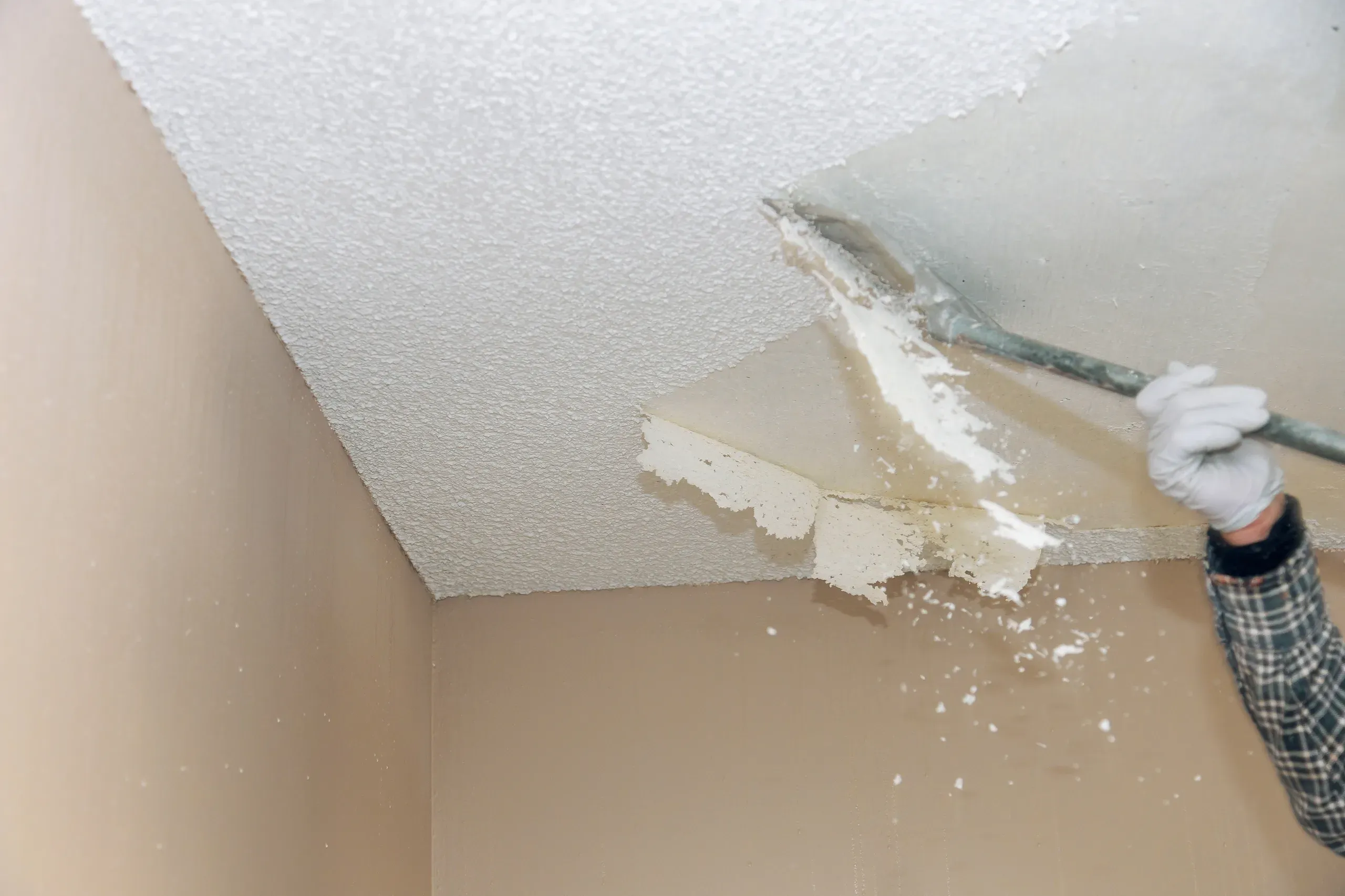 Gloved hand running a long-handled scraper across a popcorn ceiling in a room corner, half the ceiling stripped smooth and flakes of texture falling