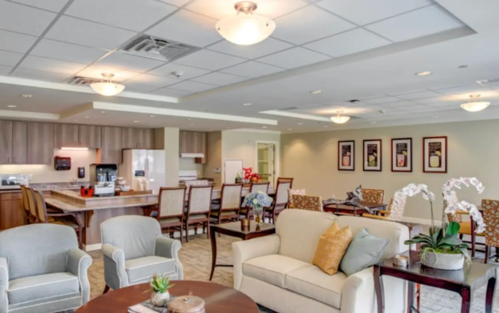 Senior-living community kitchen and dining common area with finished painted drywall, a suspended acoustic-tile ceiling with pendant fixtures, wood cabinetry, a long dining table, and lounge seating