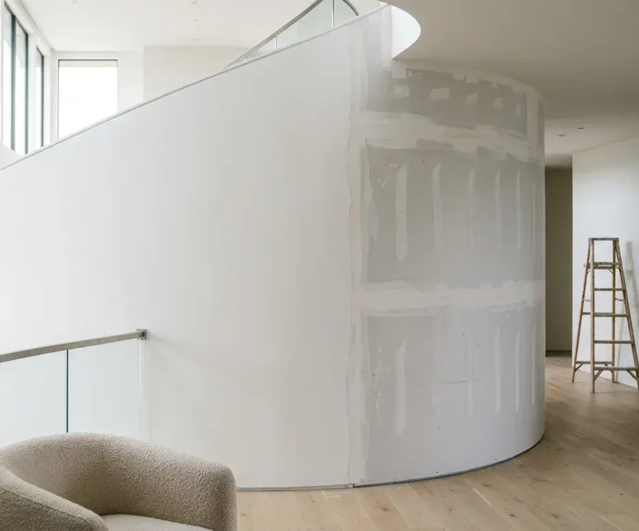 Curved interior wall being skim-coated to a Level 5 finish — one side smooth white, the other showing taped drywall joints — with a wooden ladder and a glass-railed walkway